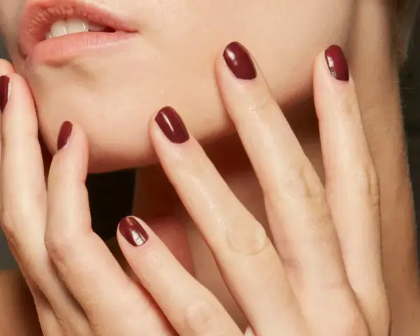 Dłonie z idealnym, ciemnoczerwonym manicurem kombinowanym. Delikatne muśnięcie policzka podkreśla elegancję.