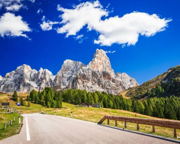 Strada panoramica per esplorare i passi del Trentino in moto, con imponenti vette dolomitiche sullo sfondo e un cielo azzurro con nuvole bianche.