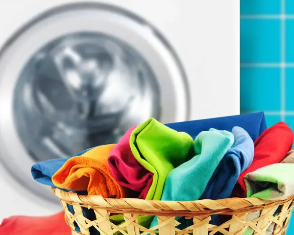 Cesto pieno di panni colorati pronti per scoprire come lavare i colorati in lavatrice. Lavatrice sullo sfondo.