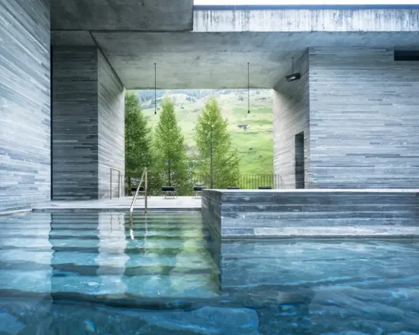 Piscine intérieure des thermes de Vals Zumthor, avec vue sur les montagnes verdoyantes et les sapins. L'eau bleue reflète les murs en pierre grise.