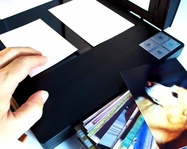 Ręka kładzie zdjęcie na skanerze, rozpoczynając proces digitalizacji zdjęć. Obok stos innych fotografii.