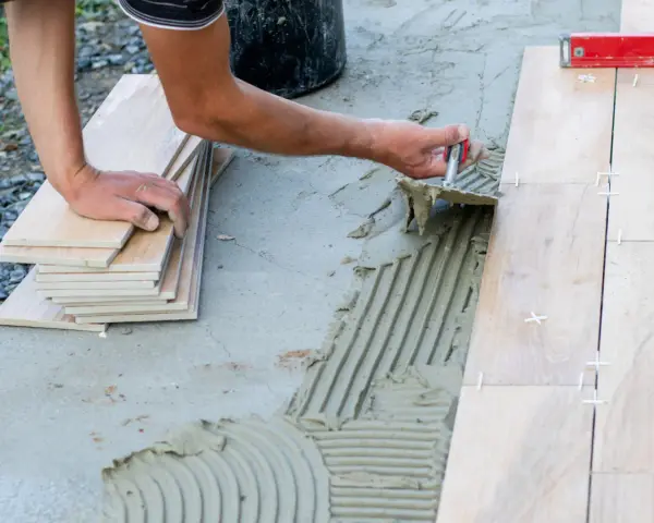 Układanie płytek na betonowym tarasie. Ręce pracownika rozprowadzają klej, obok stos płytek i poziomica.