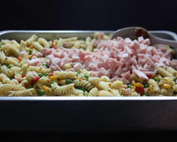 Stor ugnsform fylld med pastasallad med kassler, grönsaker och en sked.