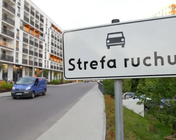 Znak "Strefa ruchu" przed nowoczesnym budynkiem mieszkalnym. Niebieski bus jedzie po drodze.
