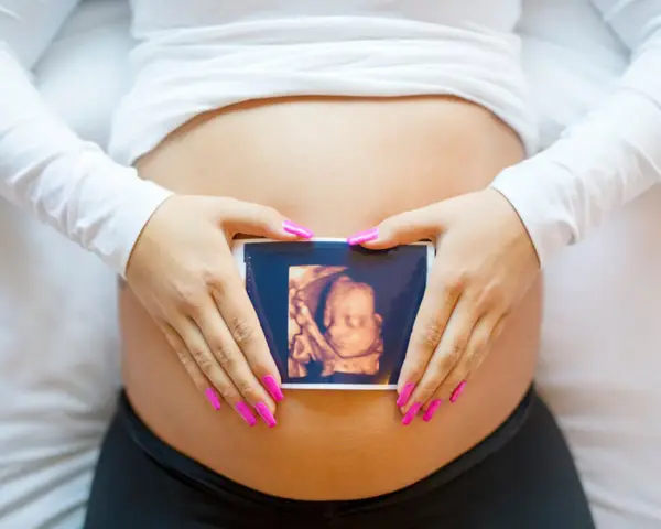 Ręce z różowymi paznokciami obejmują brzuch, na którym widnieje zdjęcie z 8 tygodnia ciąży usg 3d.
