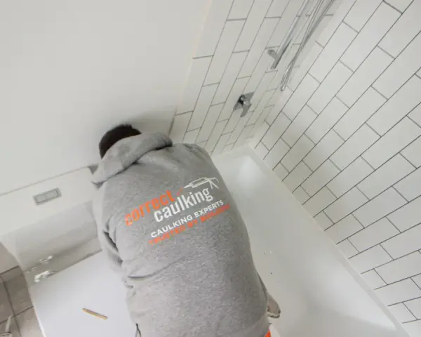 A person in a "Correct Caulking" hoodie works on a bathtub, demonstrating how to caulk a bathtub with white subway tiles in the background.