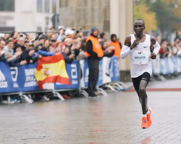 Eliud Kipchoge corre in una maratona, mostrando quanto è lunga una maratona. Pubblico festante e bandiera spagnola sullo sfondo.