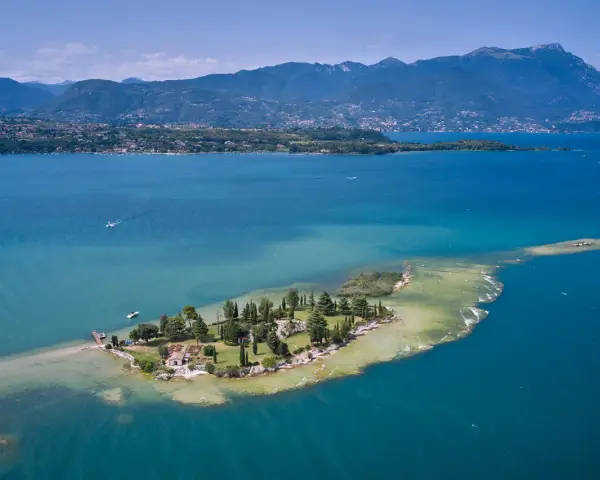 L'isola dei conigli sul Lago di Garda, un gioiello verde con case e alberi, circondata da acque turchesi e montagne sullo sfondo.
