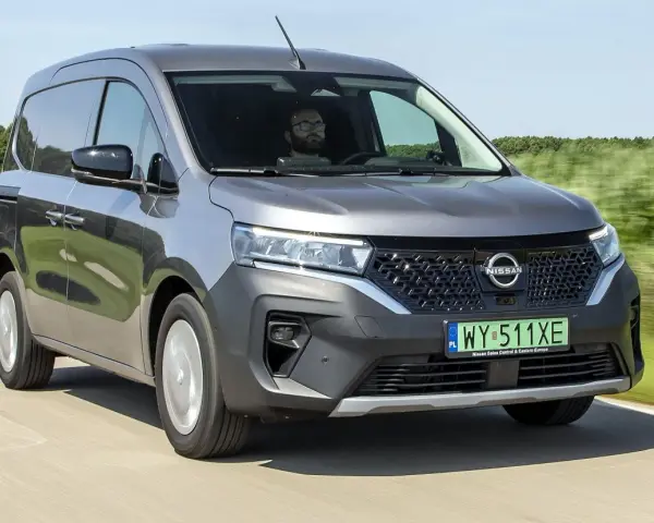 Szary Nissan Townstar jedzie po drodze, otoczony zielonymi polami. Kierowca w okularach prowadzi pojazd.