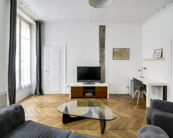 Salon moderne avec parquet en chevrons, canapés gris, bureau et TV. Les murs et le plafond sont peints à l'huile, offrant une finition impeccable.