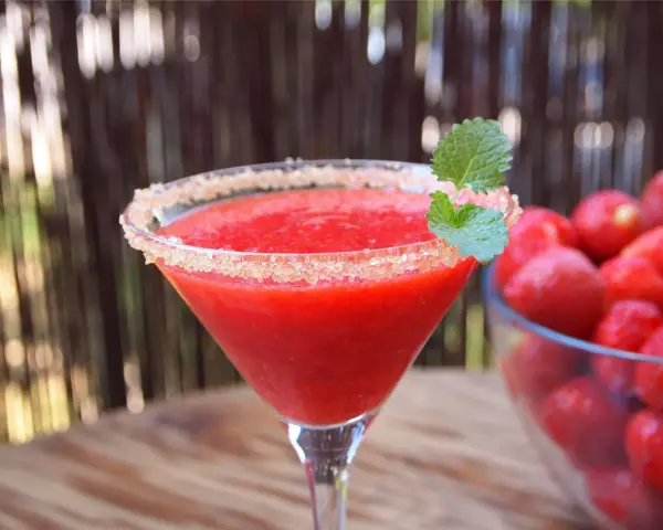 Orzeźwiający strawberry daiquiri z cukrowym rantem i listkiem mięty, obok miska pełna świeżych truskawek.
