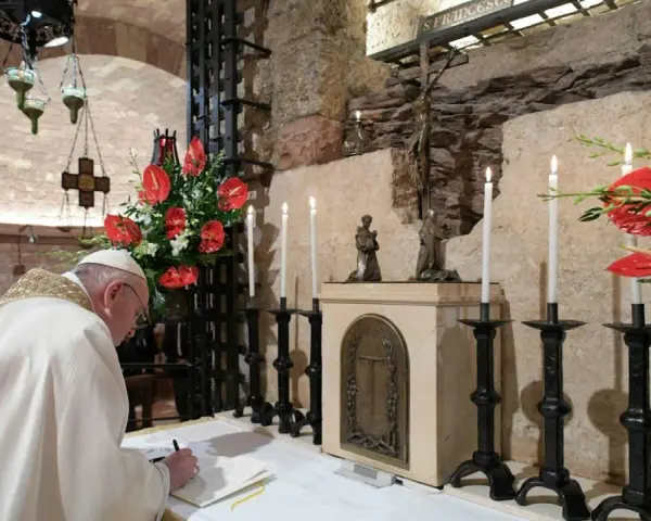 Papst Franziskus schreibt in einem Buch, während Kerzen und rote Anthurien brennen. Dies erinnert an die Bedeutung einer Enzyklika, eines wichtigen päpstlichen Schreibens.