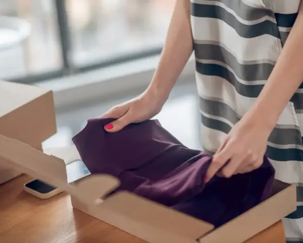 Kobieta pakuje ciemnofioletową sukienkę do kartonu, przygotowując ją do **vinted zwroty**.