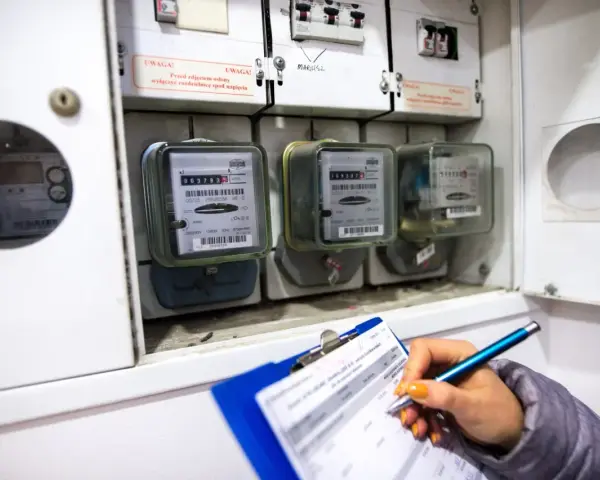 Kontrola liczników energii elektrycznej. Ręka z długopisem zapisuje dane na kartce, obok widoczne liczniki i bezpieczniki.