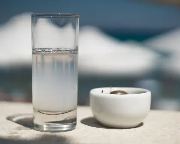 Vaso de pastis bebida, con una aceituna en un cuenco blanco. El fondo está desenfocado, sugiriendo un día soleado.
