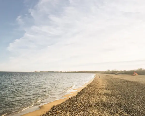 Spokojny brzeg jeziora Białe Kaszuby, fale delikatnie muskają piaszczysty brzeg, a na horyzoncie widać zarys zabudowań.