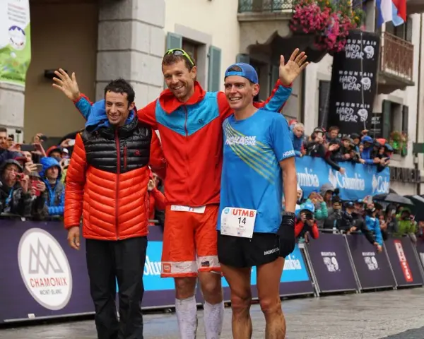 Trzech biegaczy na podium po ultra-trail du mont-blanc. Jeden w pomarańczowej kurtce, drugi w czerwonej, trzeci w niebieskiej koszulce.