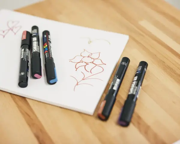 Några Posca-pennor ligger på ett vitt papper med en enkel blomma och hjärtan ritade i rött.