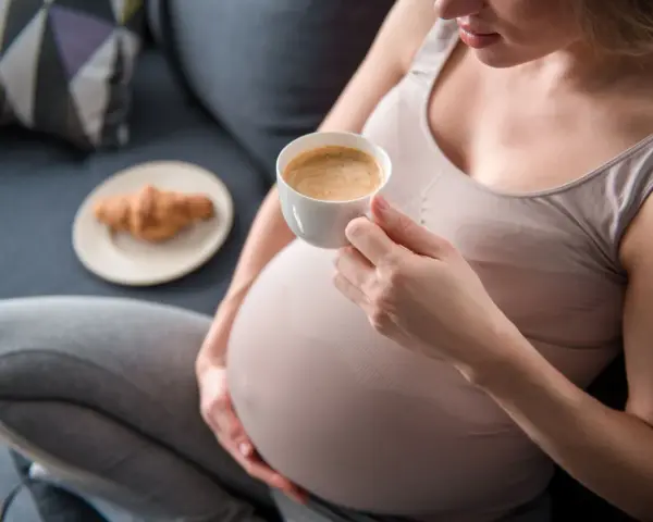Kawa w ciąży, 1 trymestr. Kobieta w ciąży pije kawę, obok leży rogalik.