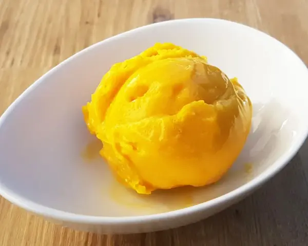Ein leuchtend gelber Löffel Mango Sorbet in einer weißen Schale auf Holzhintergrund.