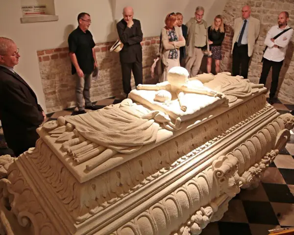 Grupa zwiedzających ogląda kamienny sarkofag w wawel krypty. Kamienne rzeźbienia zdobią jego bok.