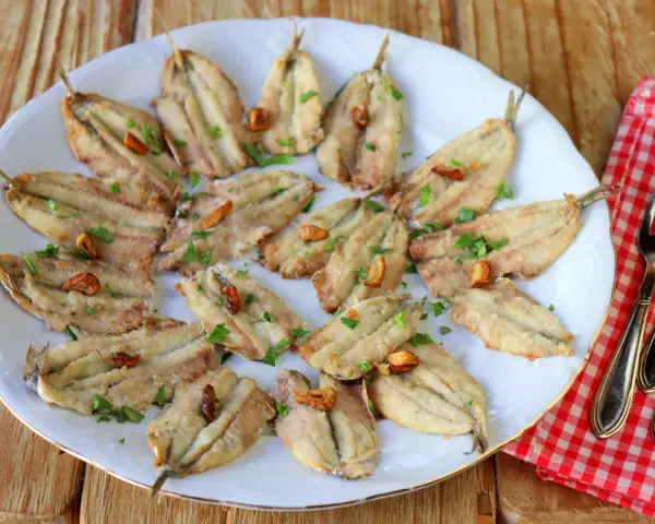 Un plato blanco con boquerones fritos, dorados y crujientes, espolvoreados con perejil fresco y trocitos de ajo.