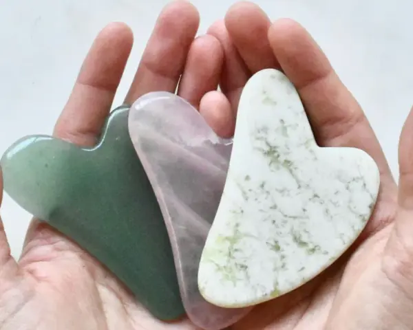 Drei herzförmige Gua Sha Steine in Grün, Rosa und Weiß liegen in einer Hand. Ideal für Gua Sha gegen Falten.