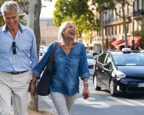 Uśmiechnięta para seniorów spaceruje po mieście z walizką, obok przejeżdżają samochody.