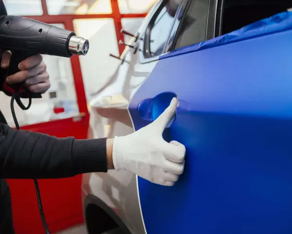 Ein Handwerker erwärmt mit einem Heißluftföhn eine blaue Folie, die auf ein Auto aufgebracht wird. Die auto folieren kosten sind hierbei ein wichtiger Faktor.