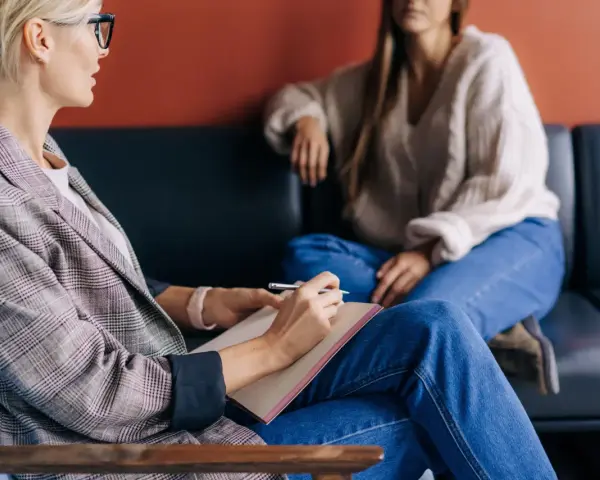 En coach jobbar med en klient. Kvinnan med glasögon skriver i en anteckningsbok medan klienten sitter avslappnad i soffan.