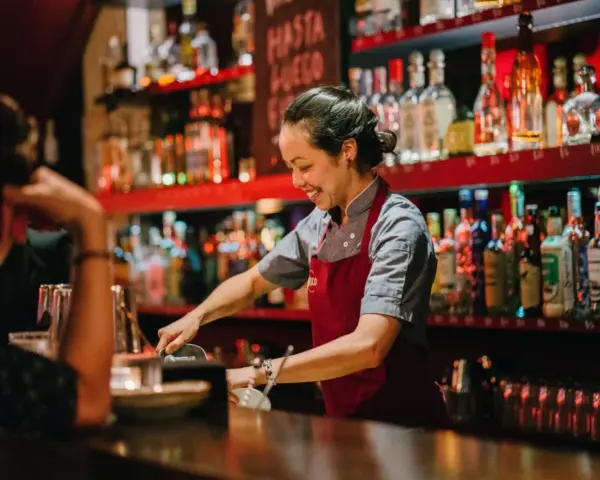 Uśmiechnięta barmanka przygotowuje drinki za barem pełnym alkoholi. Zastanawiasz się, ile zarabia barman w restauracji?