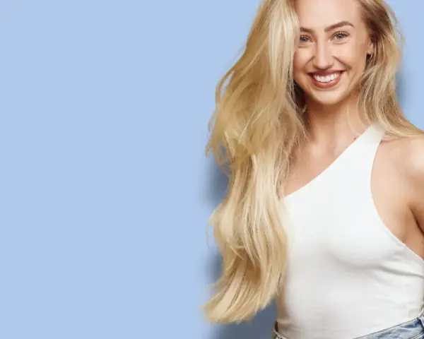 En leende kvinna med långt, blont hår som ser ut att bleka håret naturligt. Hon bär en vit topp och jeans mot en ljusblå bakgrund.