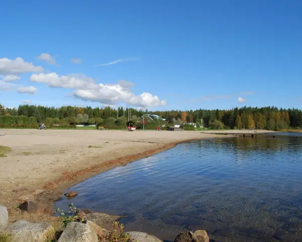 Solig strand vid Nydala camping Umeå med klart vatten och grönskande skog i bakgrunden.