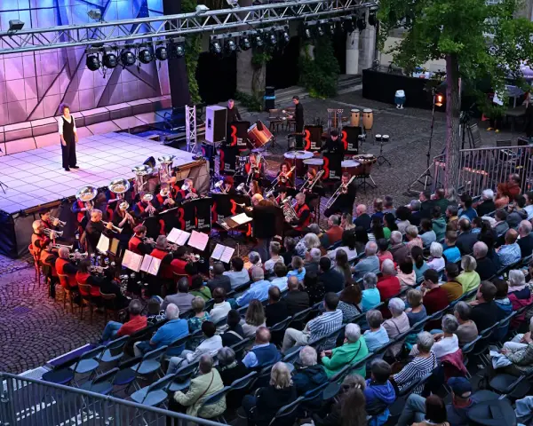 Eine Brass Band spielt auf einer Bühne, während ein Publikum auf Stühlen sitzt und zuhört.