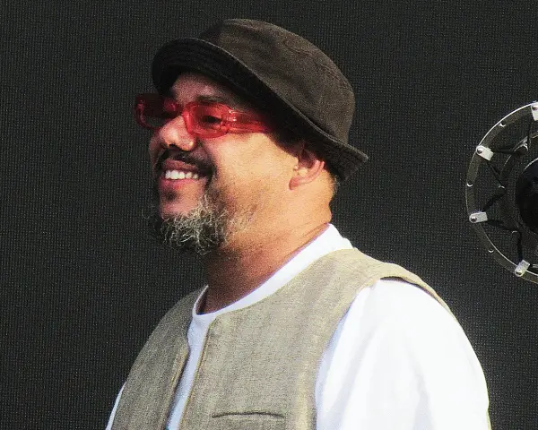 Un chanteur soul souriant, portant un chapeau et des lunettes rouges, se tient devant un microphone.