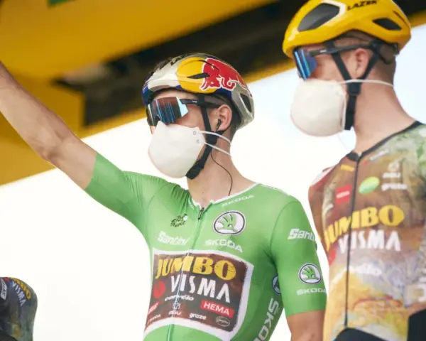 Cyclist in a green jersey, the symbol of the points classification leader at the Tour de France, raises his arm. He wears a mask and helmet.
