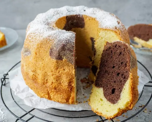 Saftiger Marmorkuchen, perfekt gebacken mit Ober-/Unterhitze oder Umluft, bestäubt mit Puderzucker. Ein Stück ist bereits angeschnitten.