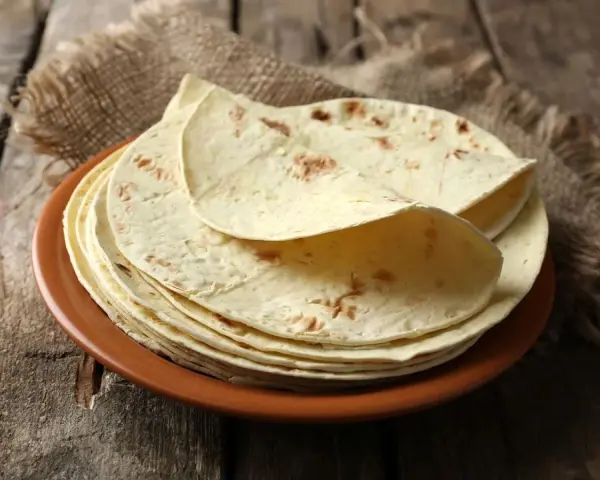 Stos cienkich, jasnych placków keto tortilla na brązowym talerzu, ułożonych na płótnie i drewnianym blacie.