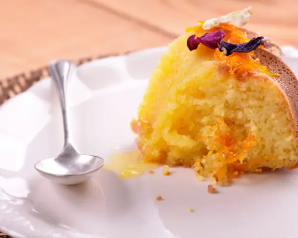 Un trozo de bizcocho de almendra de la abuela, decorado con ralladura de naranja y flores secas, servido en un plato blanco con una cucharilla.