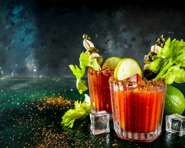 Dwa drinki Krwawa Mary z łodygami selera, limonką i piklami, ozdobione przyprawami i lodem.