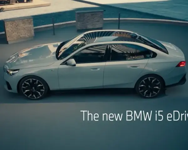 Jasnoszary sedan BMW i5 eDrive40 stoi na zewnątrz, z widokiem na basen i ławkę.