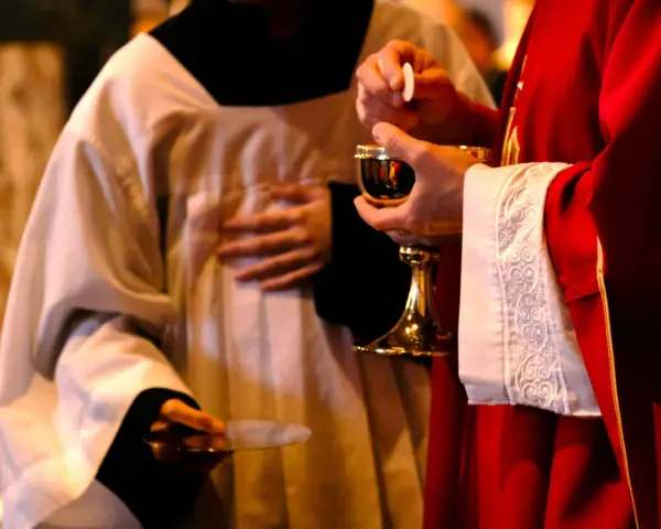 A priest in red vestments holds a chalice and host, while another in white vestments holds a paten. A moment of communion, perhaps a prayer to St. Benedict for healing.