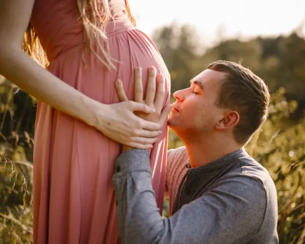 Ciepła sesja ciążowa z mężem. On klęczy i całuje brzuszek ukochanej, otoczeni złotym światłem i naturą.