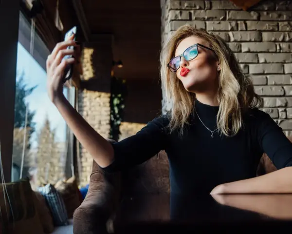 Kobieta w okularach robi sobie selfie, wyciągając usta do pocałunku. W jej spojrzeniu widać pewność siebie, graniczącą z narcyzmem.