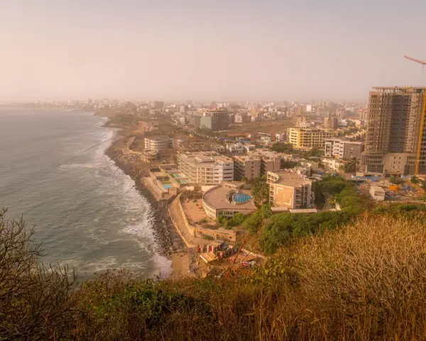 Widok na wybrzeże i rozwijające się miasto w **Afryce, Senegal**. Widać ocean, plażę, nowoczesne budynki i plac budowy z dźwigiem.