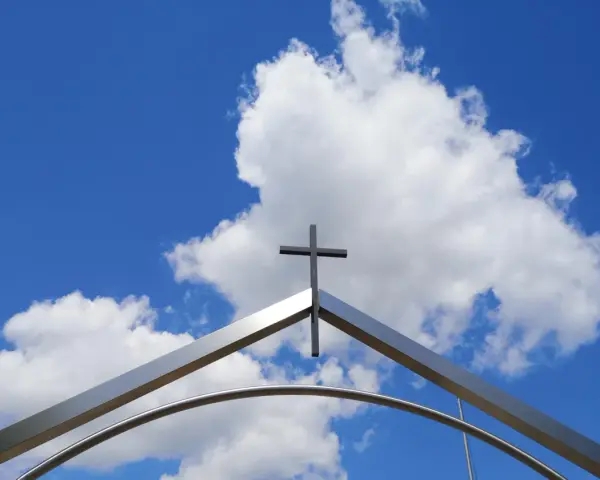 Ein Kreuz ragt über einem modernen Torbogen in den blauen Himmel mit Wolken. Ein starkes Christi Himmelfahrt Symbol.