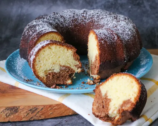 Saftiger weltbester Marmorkuchen, mit Puderzucker bestäubt, auf blauem Teller. Ein Stück ist abgetrennt und zeigt die köstliche Marmorierung.