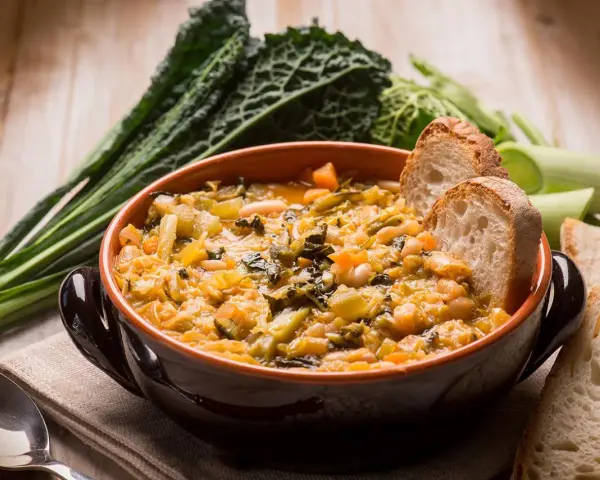 Una ciotola di ribollita fumante, ricca di verdure e fagioli, accompagnata da fette di pane tostato e cavolo nero.