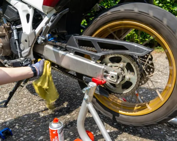 Mano guantata pulisce la catena della moto con uno straccio giallo. Spray per la pulizia catena visibile.