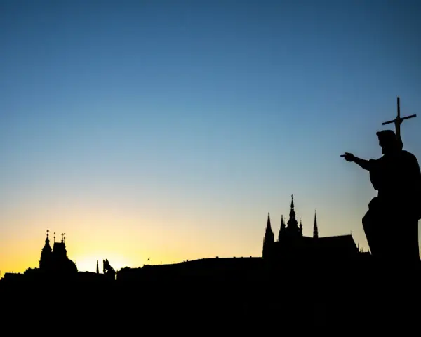 Silhouette d'une statue de saint pointant vers un paysage urbain au coucher du soleil. Le ciel bleu dégradé est le centre d'intérêt de l'image.